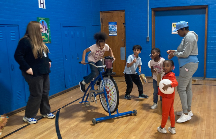  a student on a bike powering a blender to make a blender surrounded by some adults and other studen